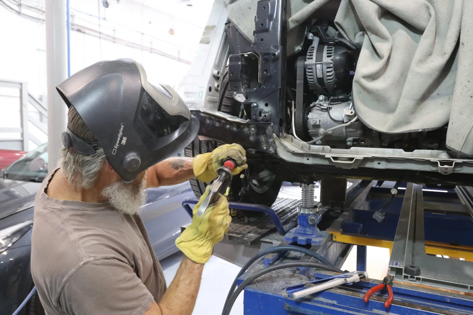 A technician wearing a welding helmet and gloves preps the frame of a vehicle for welding in an automotive repair shop.