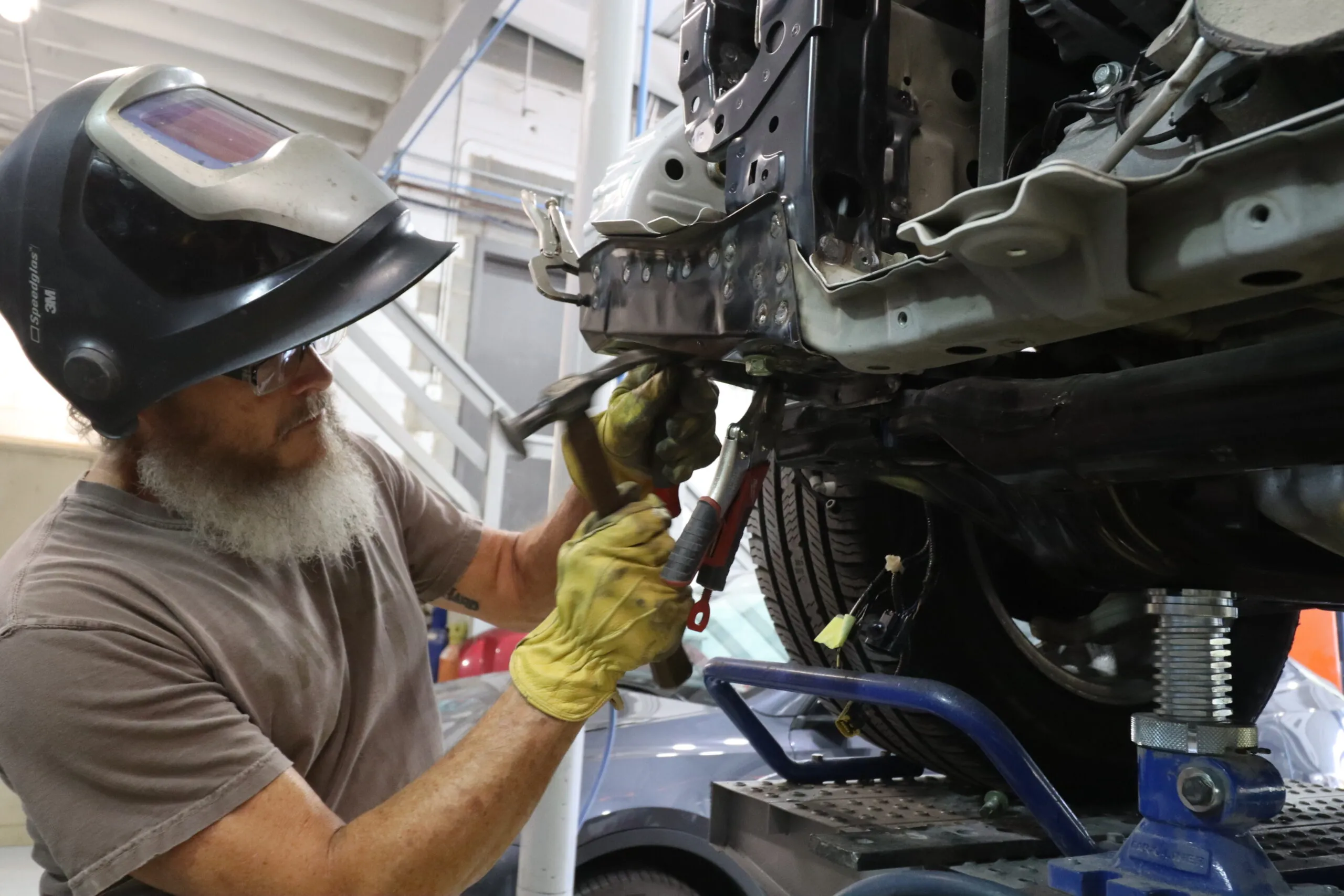 A technician wearing a welding helmet and gloves works on the frame of a vehicle on an elevated platform in an automotive repair shop.