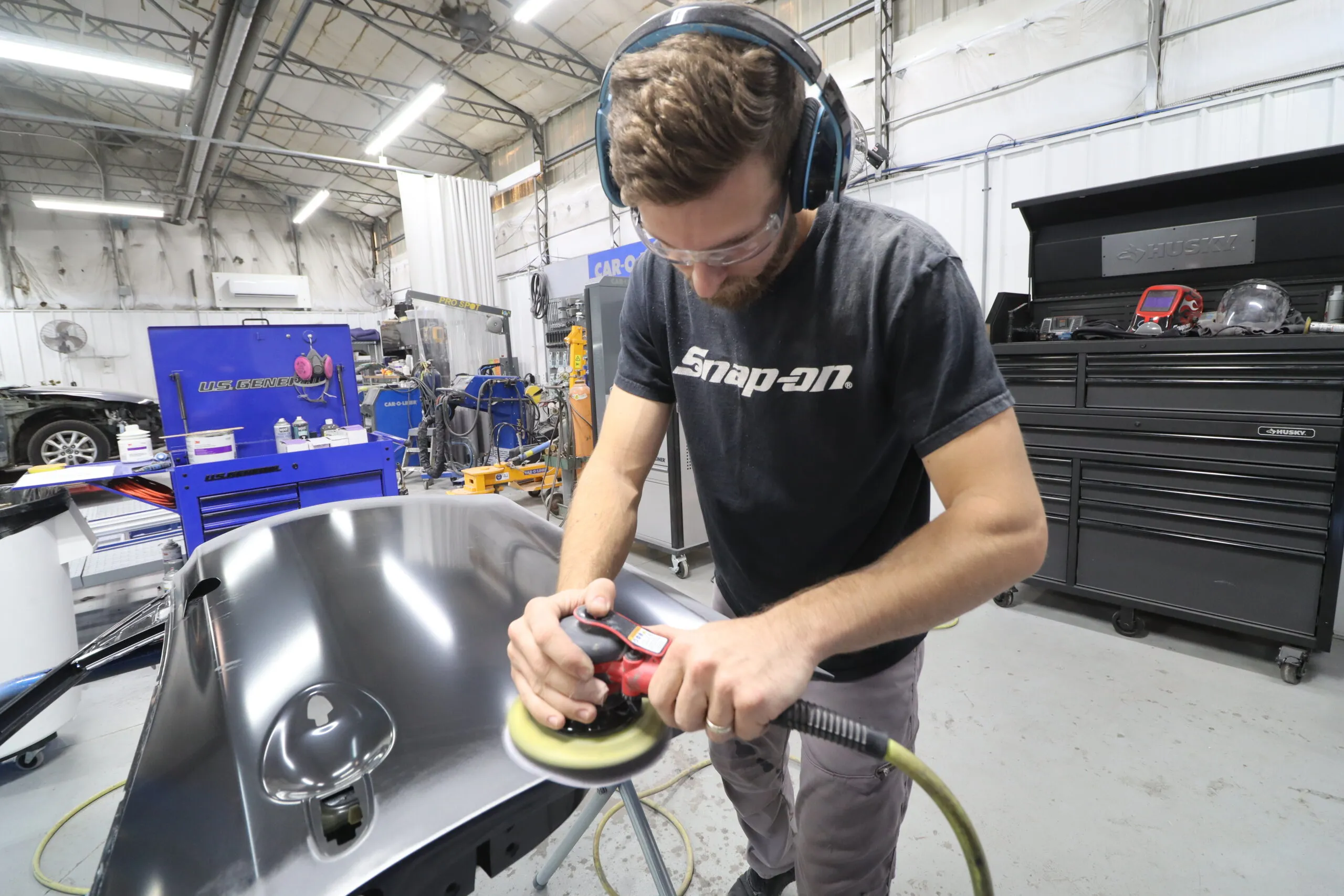 A technician wearing safety goggles and headphones polishes a car panel with a sanding tool in an auto body repair shop.