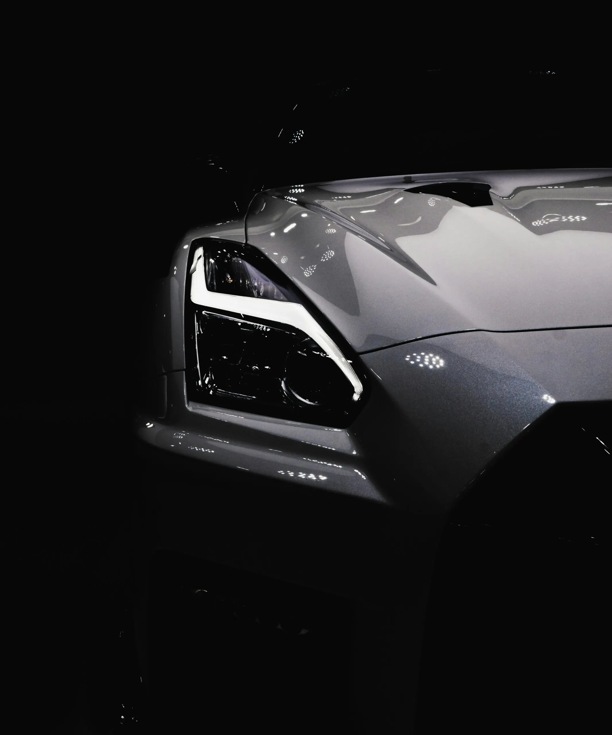 Front close-up of a sleek gray Nissan with illuminated headlights in a dark background.