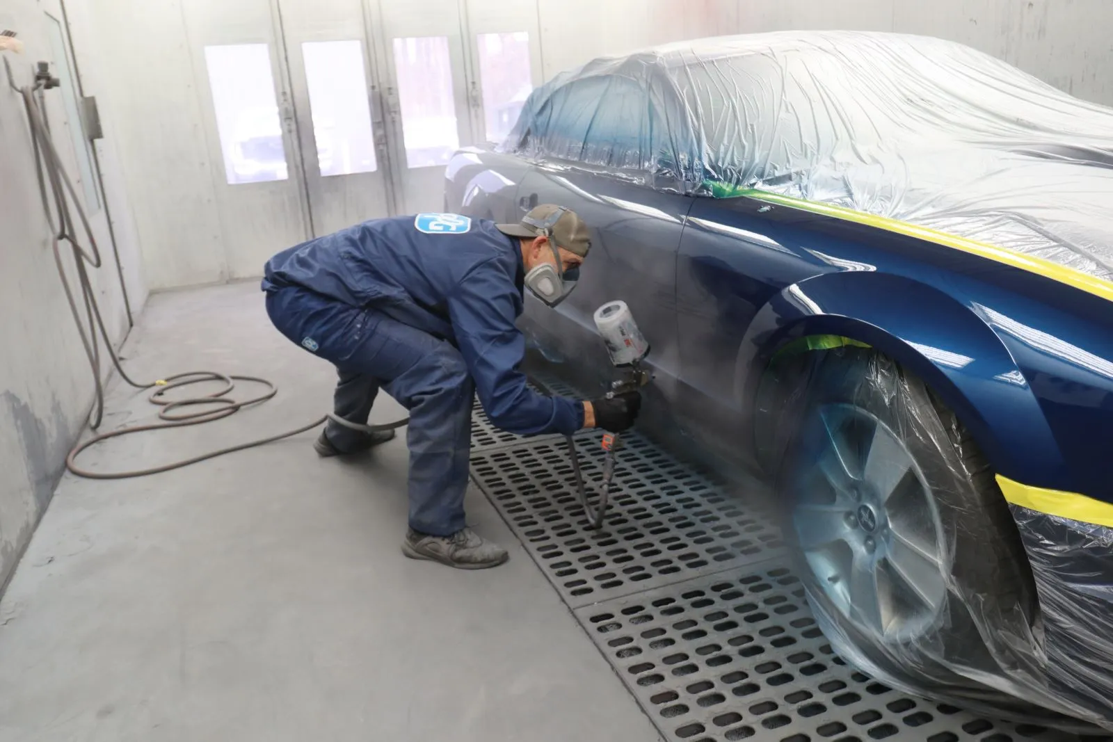 Automotive technician wearing safety gear while using a paint spray gun to refinish a vehicle inside a clean paint booth.