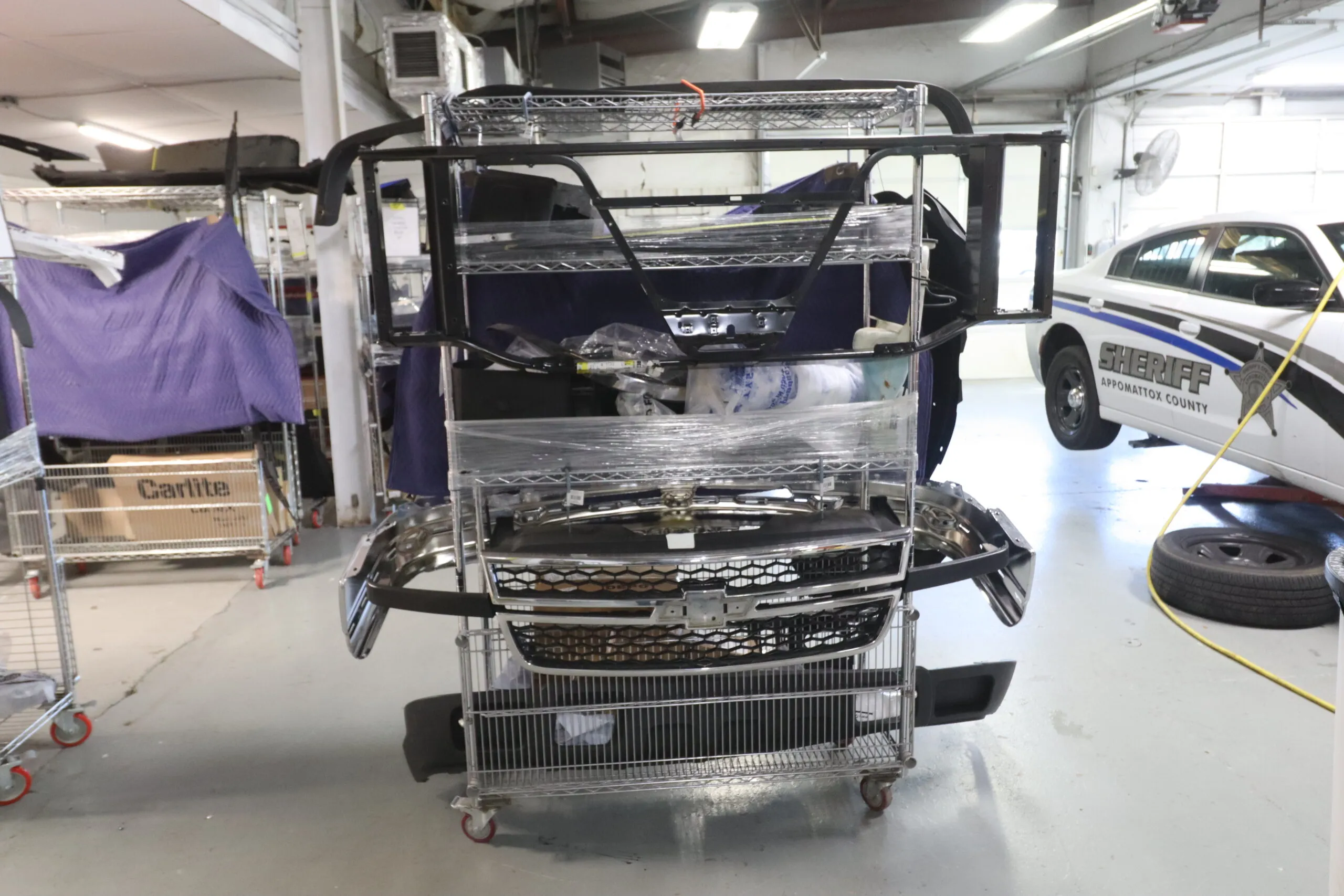 Organized automotive parts on a cart at Sandrof Auto Body repair shop, with a Sheriff’s vehicle in the background.