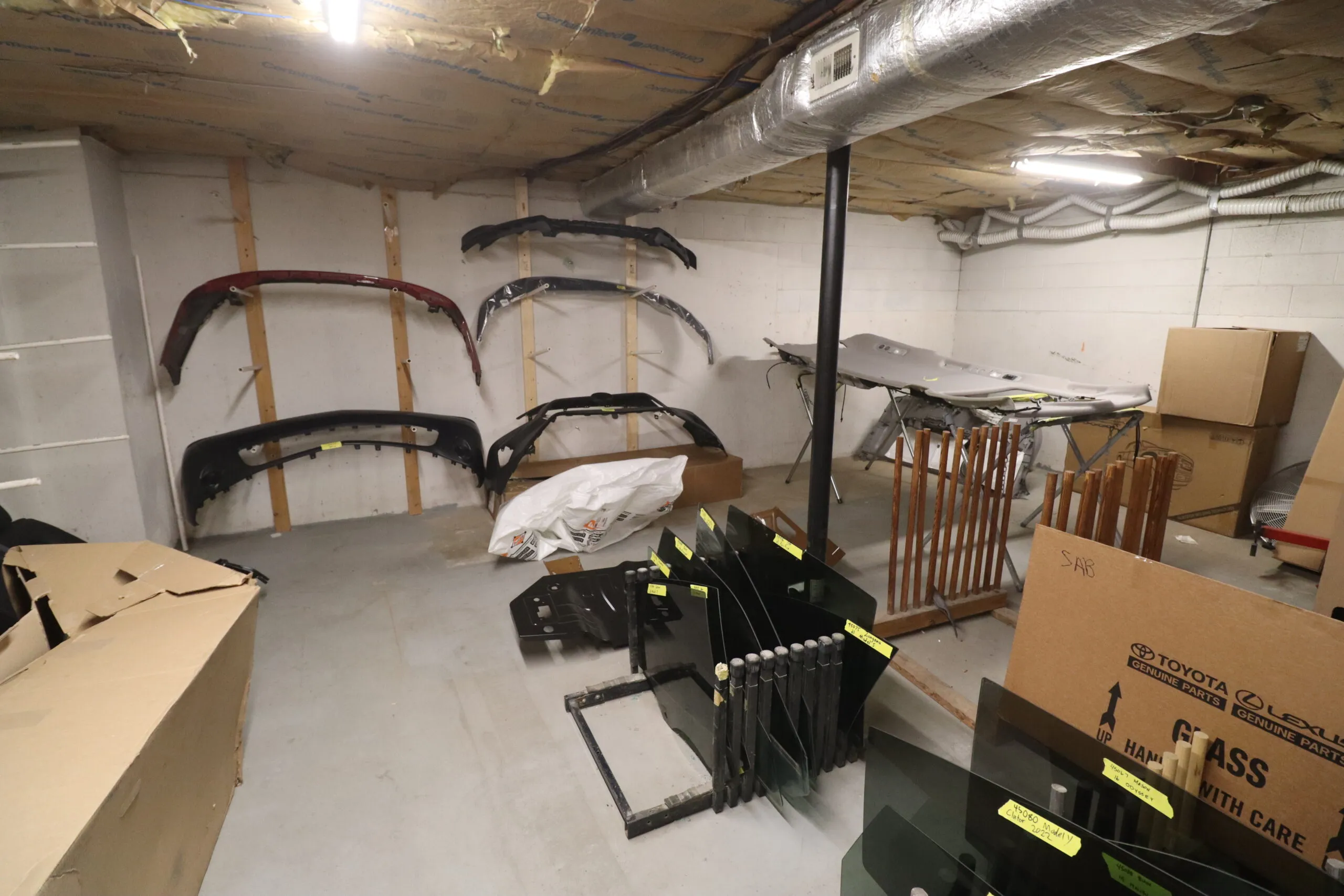 A storage area in a body shop with vehicle bumpers mounted on the wall, auto glass panels stacked in frames, and other car parts organized across the space.