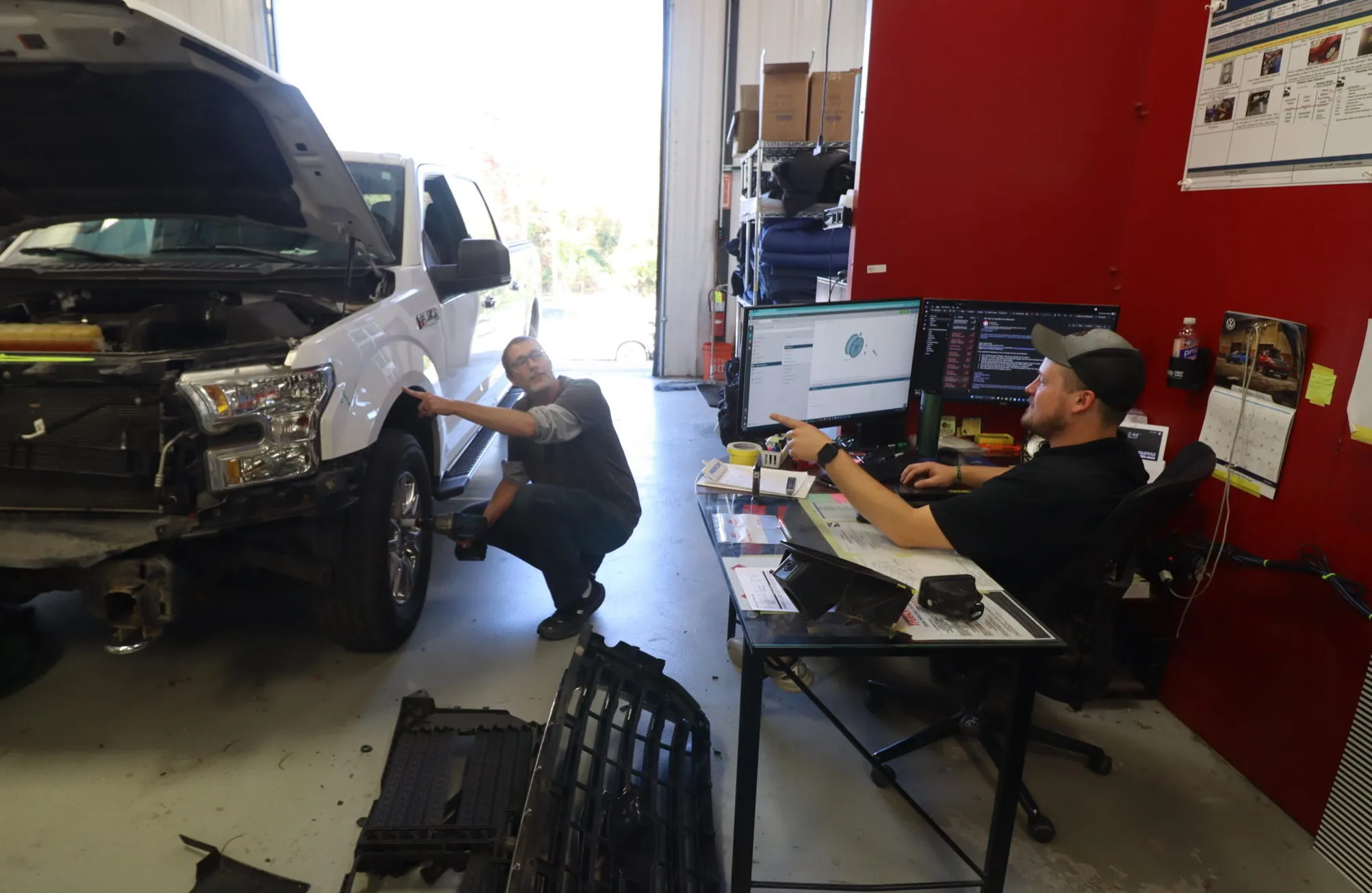 A technician working on a white truck's wheel assembly while a repair planner at a desk reviews digital schematics at Sandrof Auto Body on his Computer.